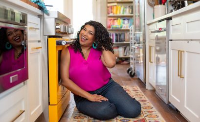 aarti sequeira wears her curly hair while sitting on the floor and smiling in front of her yellow kitchen oven